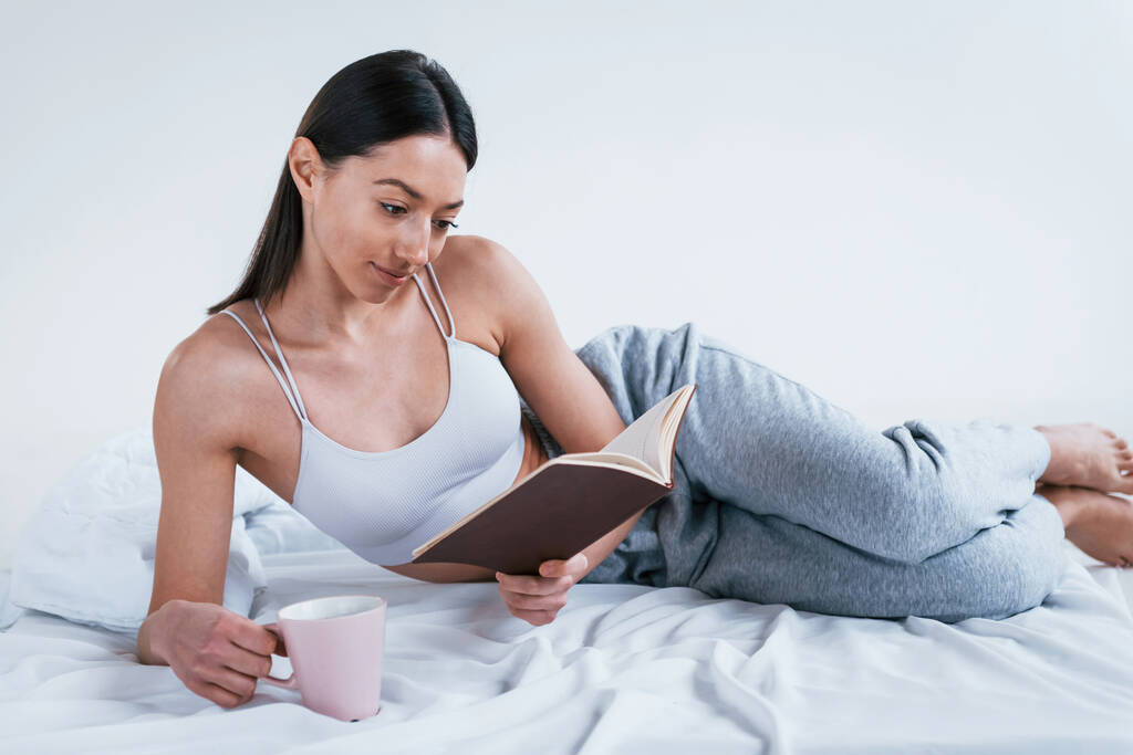 Позитивная улыбающаяся женщина в спортивной одежде лежит в студии с книгой и чашкой напитка в руках. - Фото, изображение