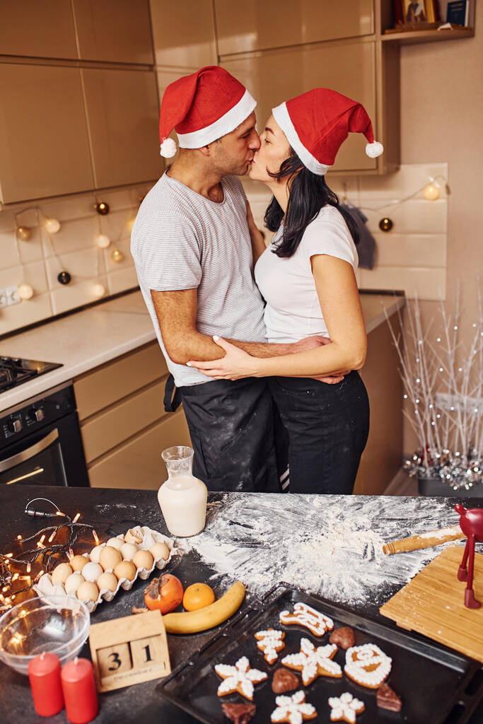 Abraçar e beijar. Casal com chapéus de Papai Noel em pé na cozinha juntos dentro de casa. - Foto, Imagem