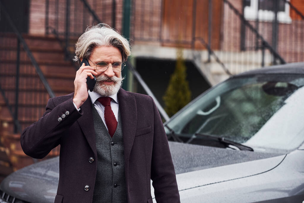 El hombre mayor de moda con el pelo gris y la barba está al aire libre en la calle cerca de su coche tener conversación por teléfono. - Foto, Imagen