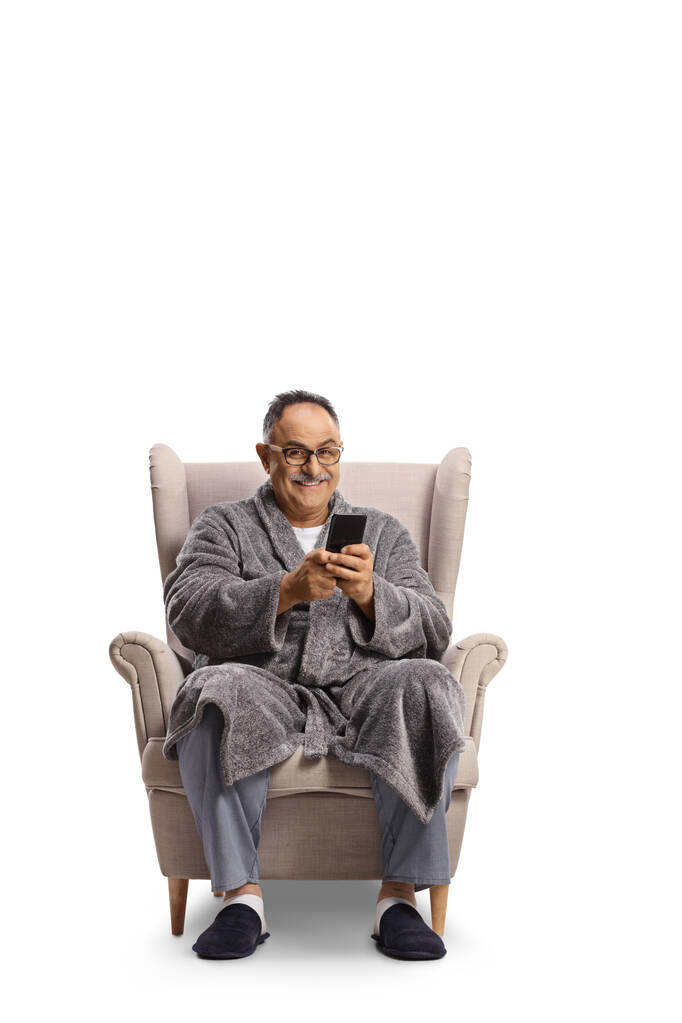 Homem maduro em um roupão de banho sentado em uma poltrona e usando um smartphone isolado no fundo branco - Foto, Imagem