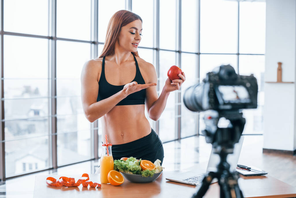 Женщина-блогер со спортивным телом, стоящая в помещении рядом со столом со здоровой пищей. - Фото, изображение