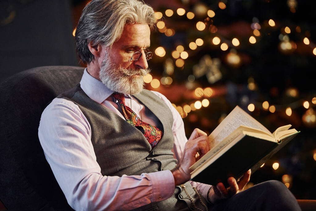 Πορτρέτο της κομψής ανώτερος με γκρι μαλλιά και γενειάδα βιβλίο ανάγνωσης σε διακοσμημένο χριστουγεννιάτικο δωμάτιο. - Φωτογραφία, εικόνα