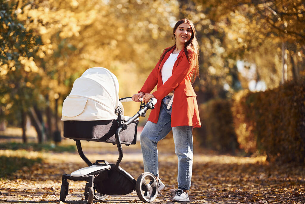 Mother in red coat have a walk with her kid in the pram in the park with beautiful trees at autumn time. - Photo, Image