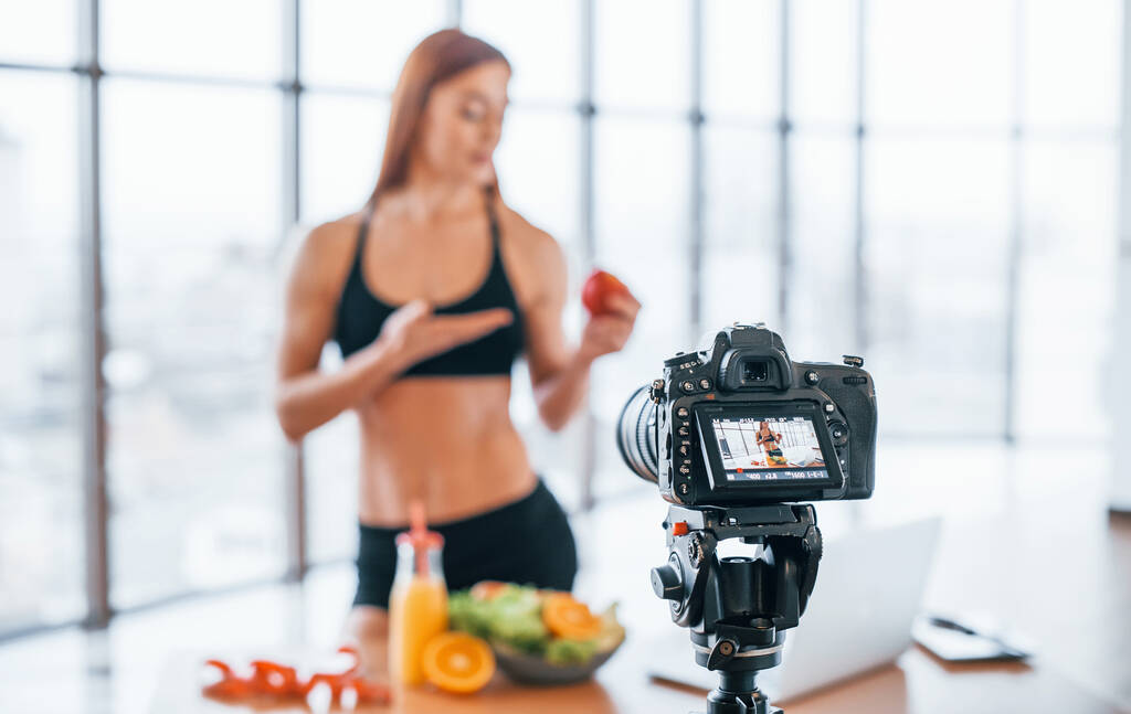 Kobieta vlogger ze sportowym ciała stojącego w pomieszczeniach w pobliżu stołu ze zdrową żywnością. - Zdjęcie, obraz