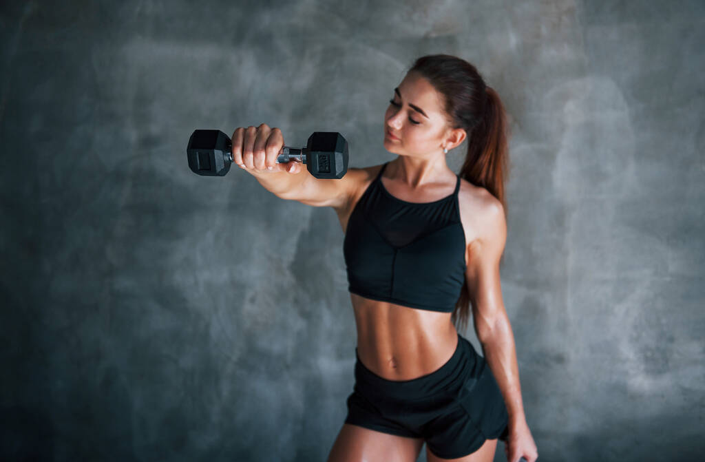 Junge Fitness-Frau steht mit Hanteln in der Hand im Fitnessstudio an der Wand. - Foto, Bild