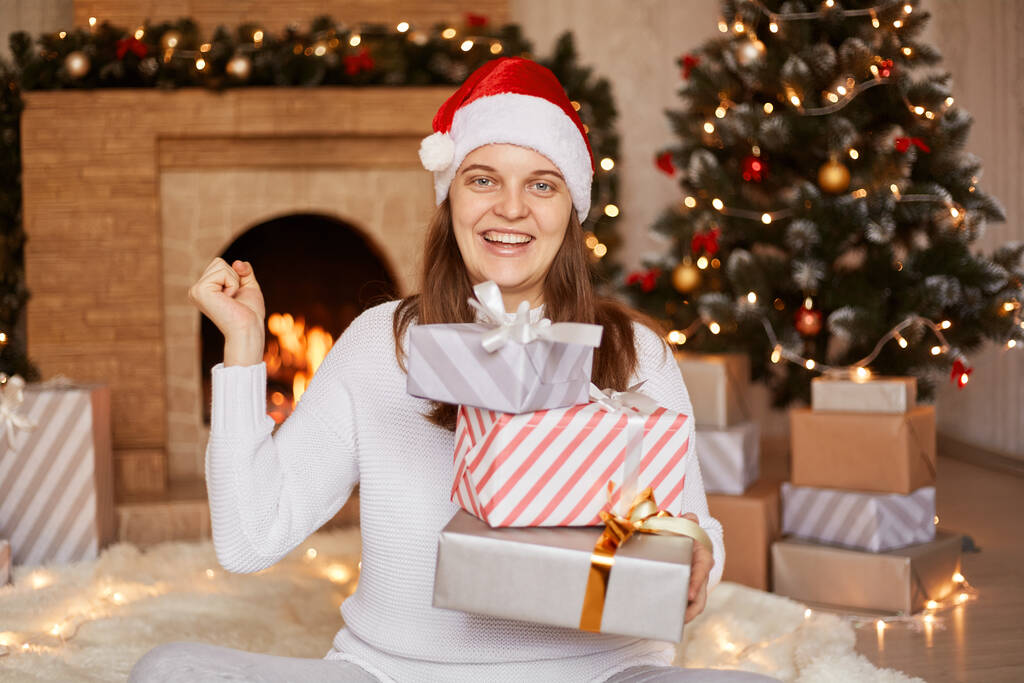 Εσωτερική λήψη χαρούμενης ενθουσιασμένης γυναίκας με λευκό πουλόβερ και καπέλο santa claus, να είναι σε εορταστικό δωμάτιο με τζάκι και χριστουγεννιάτικο δέντρο, να κάθεται με τη στοίβα των δώρων του νέου έτους. - Φωτογραφία, εικόνα