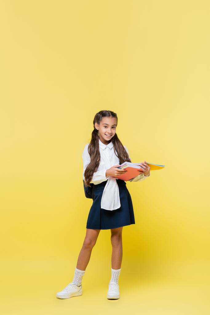 Vrolijk schoolmeisje met notitieboekjes en kijken naar camera op gele achtergrond  - Foto, afbeelding