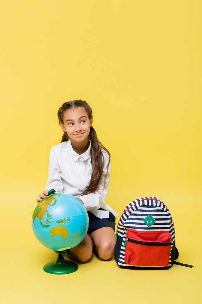Escolar sonriente sentado cerca de globo y mochila sobre fondo amarillo  - Foto, Imagen