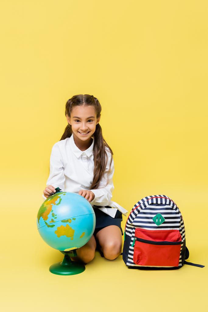 Счастливый ребенок смотрит на камеру рядом с глобусом и рюкзаком на желтом фоне  - Фото, изображение