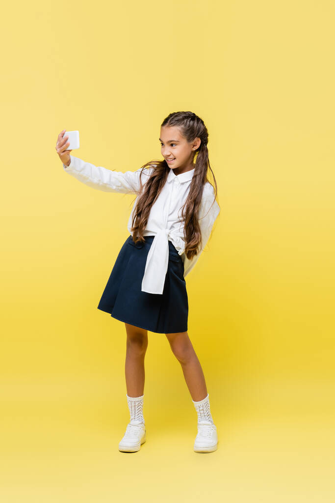 Gülümseyen okul çocuğu sarı arka planda cep telefonuyla selfie çekiyor.  - Fotoğraf, Görsel