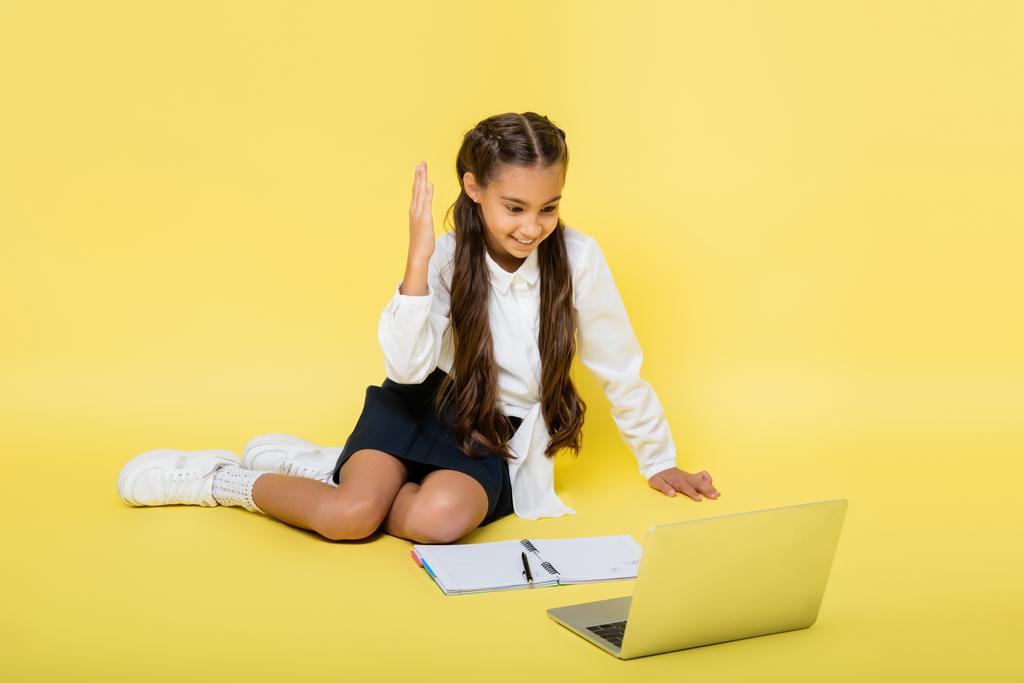 Улыбающийся школьник поднимает руку во время видеозвонка на ноутбук рядом с ноутбуком на желтом фоне - Фото, изображение
