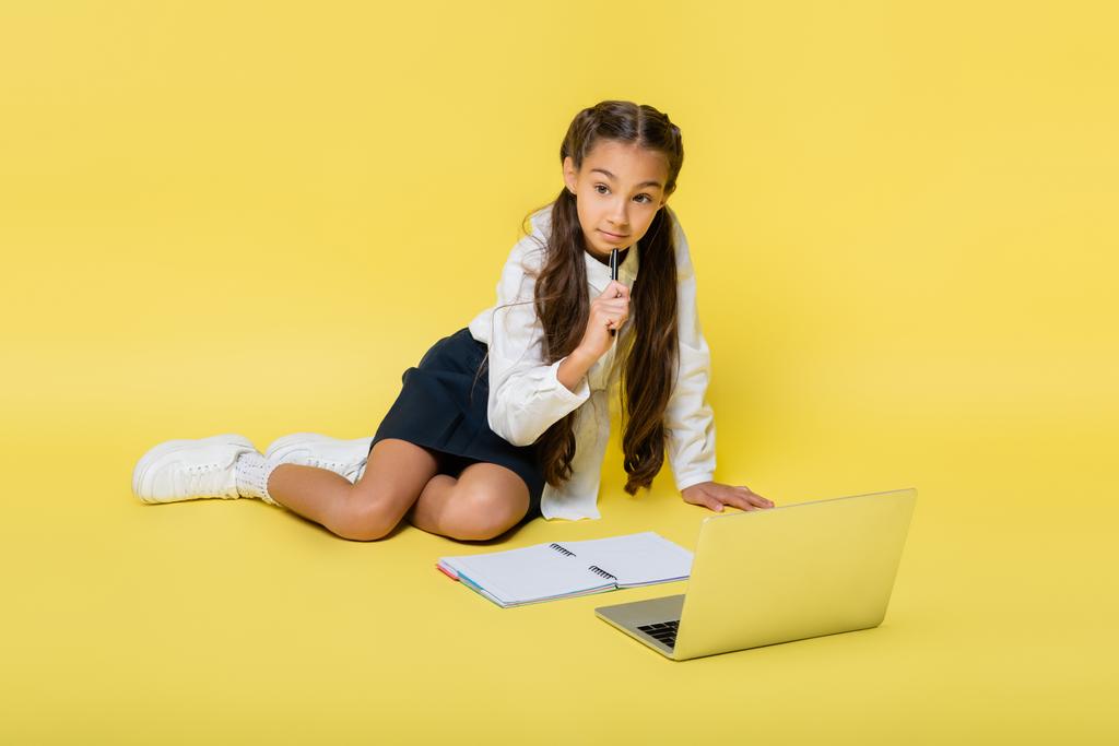 Pensive schoolkid holding pen near laptop and copy book on yellow background - Photo, Image