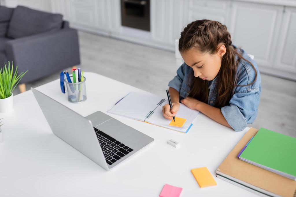 Bambino preadolescente che scrive sulla nota appiccicosa sul notebook vicino al laptop a casa  - Foto, immagini