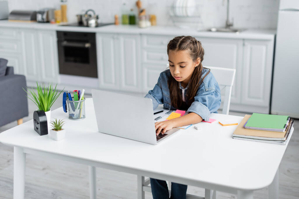 Preteen enfant en utilisant un ordinateur portable pendant l'éducation en ligne près de copier des livres et des plantes à la maison  - Photo, image