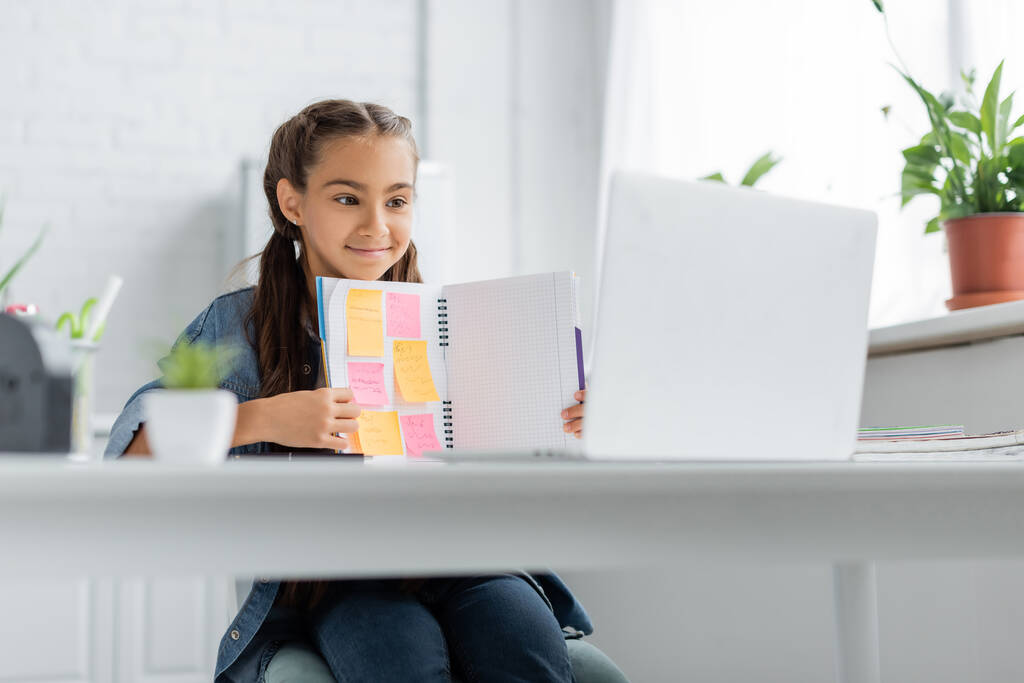 Schoolkind houdt notebook met plakkerige notities in de buurt wazig laptop tijdens videogesprek thuis  - Foto, afbeelding