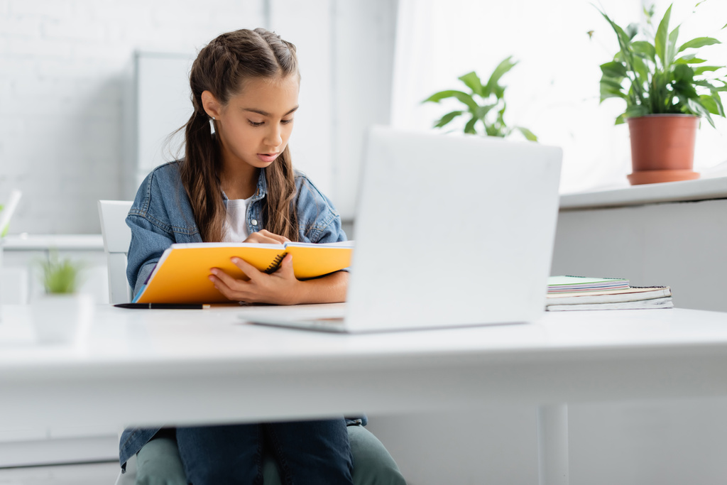 Chica preadolescente sosteniendo portátil cerca borrosa portátil durante la educación en línea en el hogar  - Foto, imagen