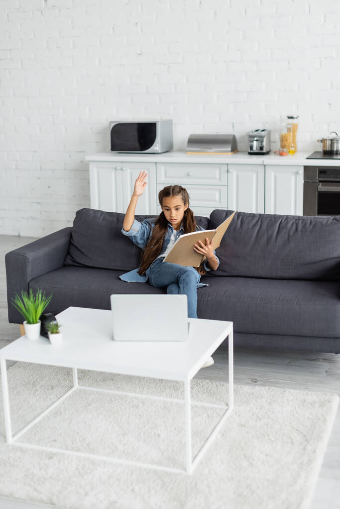 Ребенок-подросток с ноутбуком, поднимающим руку возле ноутбука на диване дома  - Фото, изображение