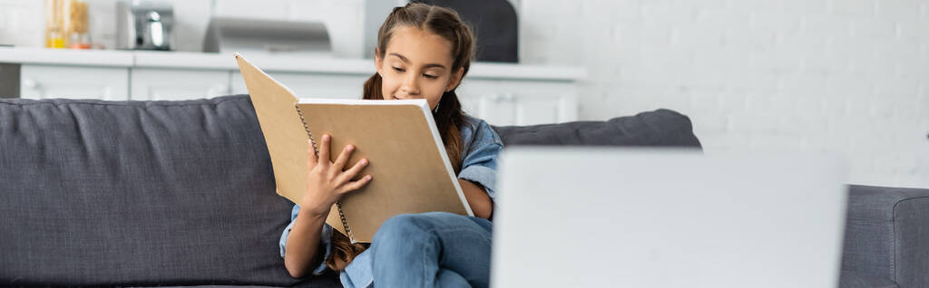 Παιδί κρατώντας σημειωματάριο κοντά θολή φορητό υπολογιστή στο σπίτι, πανό  - Φωτογραφία, εικόνα