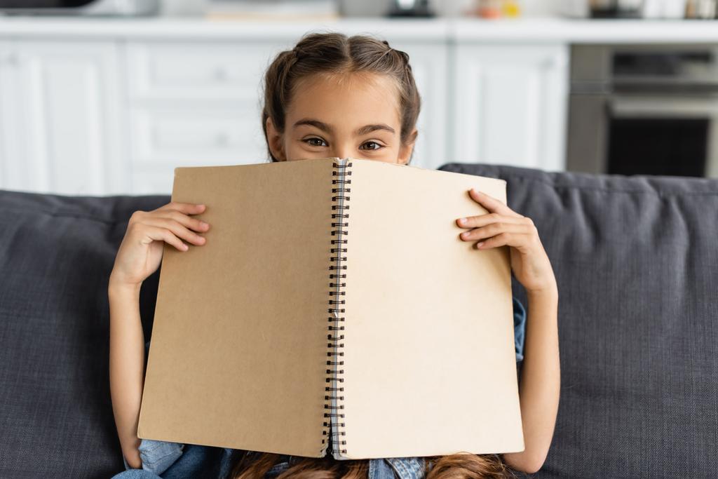 Preteen kid covering face with notebook and looking at camera on couch at home  - Photo, Image