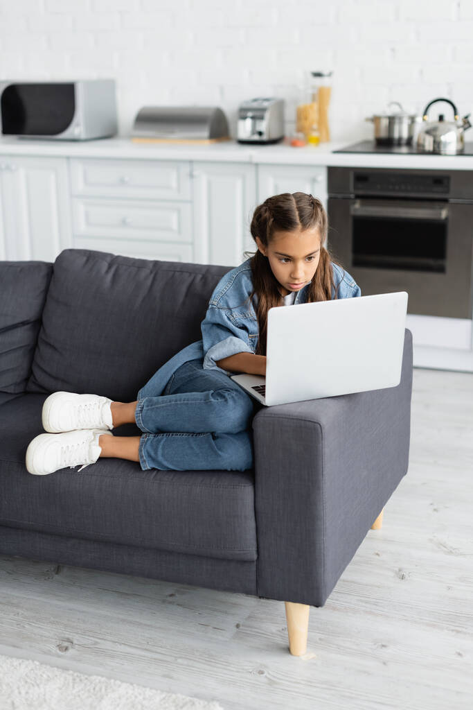 Niño preadolescente usando el ordenador portátil mientras está sentado en el sofá en casa  - Foto, imagen