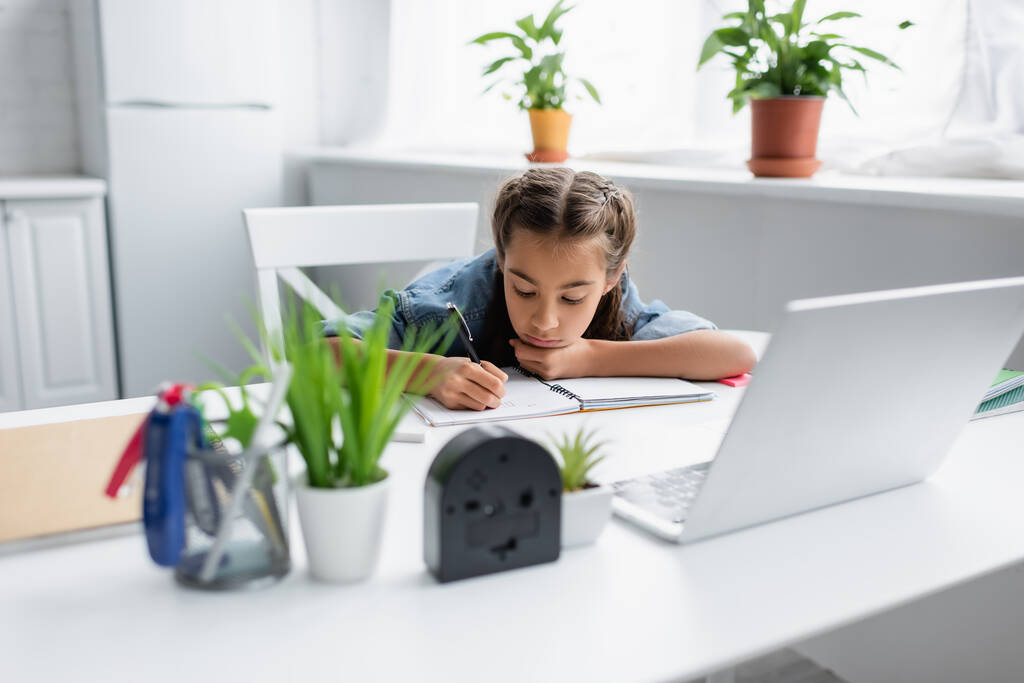 Criança escrevendo em livro de cópia perto de laptop borrado e plantas em casa  - Foto, Imagem