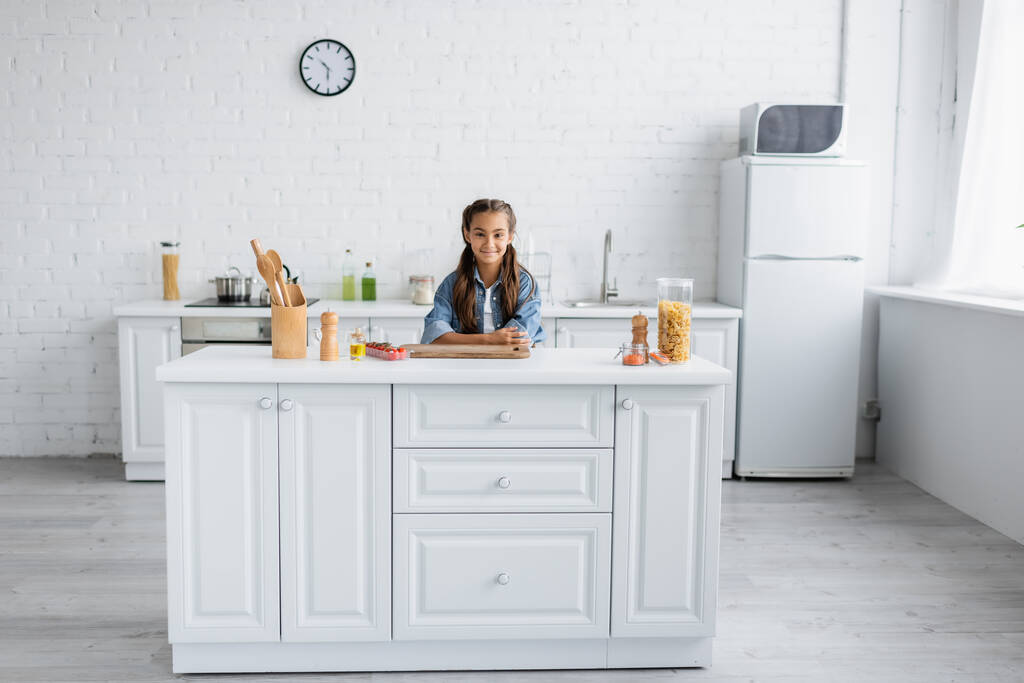 Lächelndes Kind blickt in die Kamera neben Makkaroni und Kirschtomaten in der Küche  - Foto, Bild