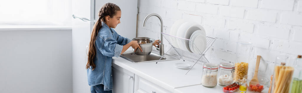 Πλάγια άποψη του παιδιού ρίχνει νερό στο κατσαρολάκι κοντά στο φαγητό στην κουζίνα, πανό  - Φωτογραφία, εικόνα