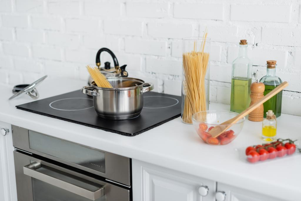 Macaroni near cherry tomatoes and saucepan on stove in kitchen  - Photo, Image