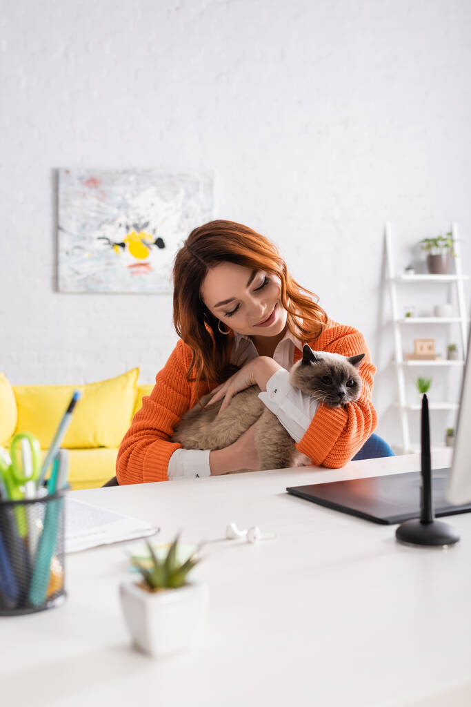 счастливая женщина, обнимающая кота, сидя за столом рядом с размытым графическим планшетом и стилусом - Фото, изображение