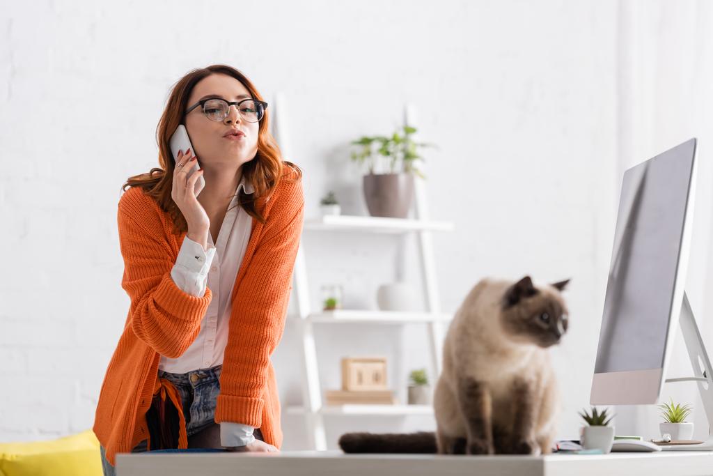 nő szemüveges duzzogó ajkak, miközben hívja a mobiltelefon közelében elmosódott macska az asztalon - Fotó, kép