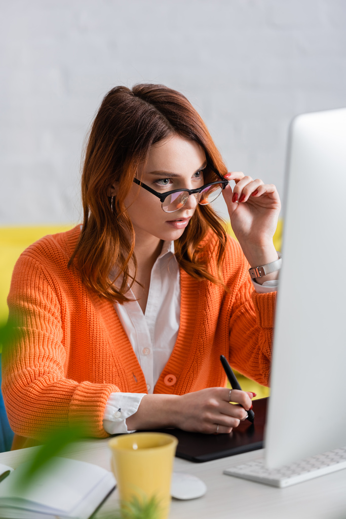freelancer concentrado tocando óculos enquanto trabalhava em tablet gráfico e olhando para monitor desfocado - Foto, Imagem