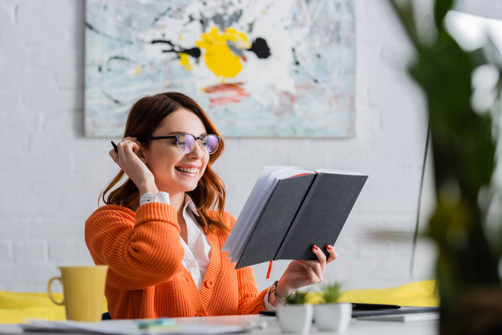 vrolijke vrouw in bril kijken naar notebook tijdens het werken thuis op wazig voorgrond - Foto, afbeelding