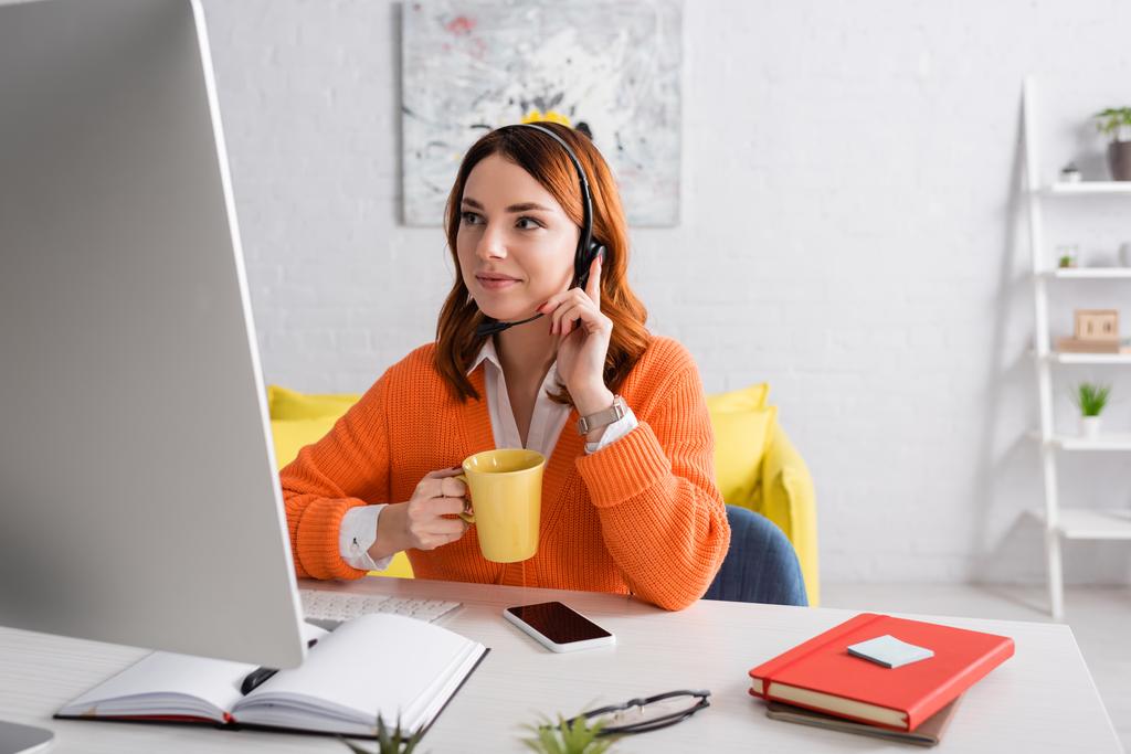 donna sorridente in cuffia che tiene una tazza di tè mentre lavora vicino al computer e allo smartphone con schermo bianco - Foto, immagini
