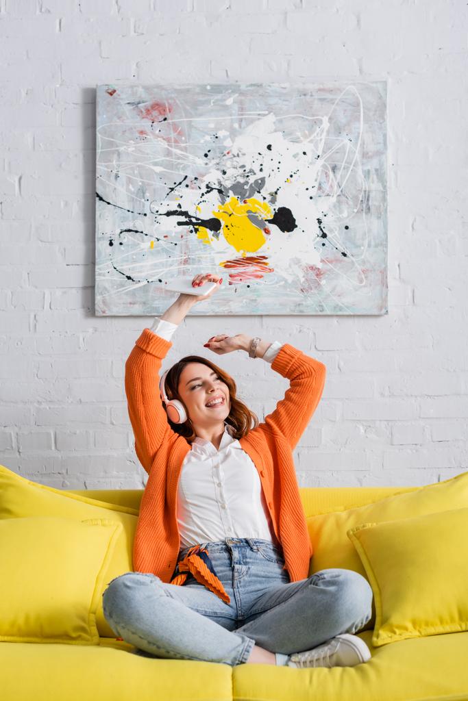 mulher alegre sentado no sofá com as mãos levantadas enquanto ouve música em fones de ouvido - Foto, Imagem