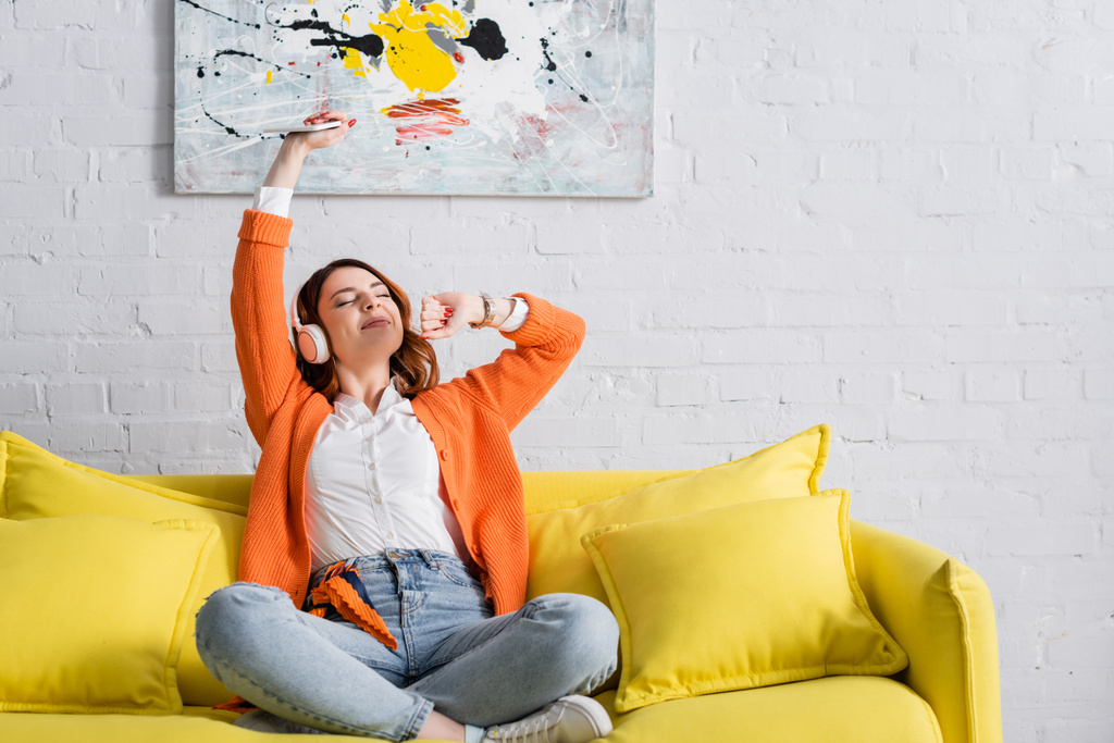 femme excitée avec smartphone dans la main levée écouter de la musique sur le canapé jaune - Photo, image