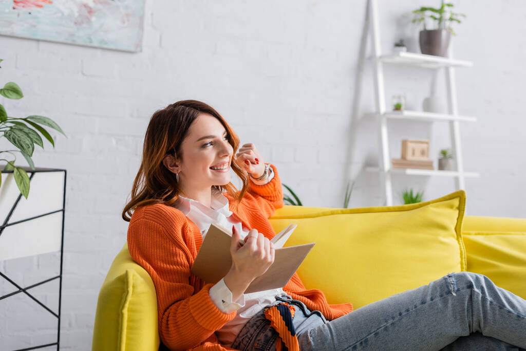 glückliche Frau mit Buch schaut weg, während sie zu Hause auf dem Sofa sitzt - Foto, Bild