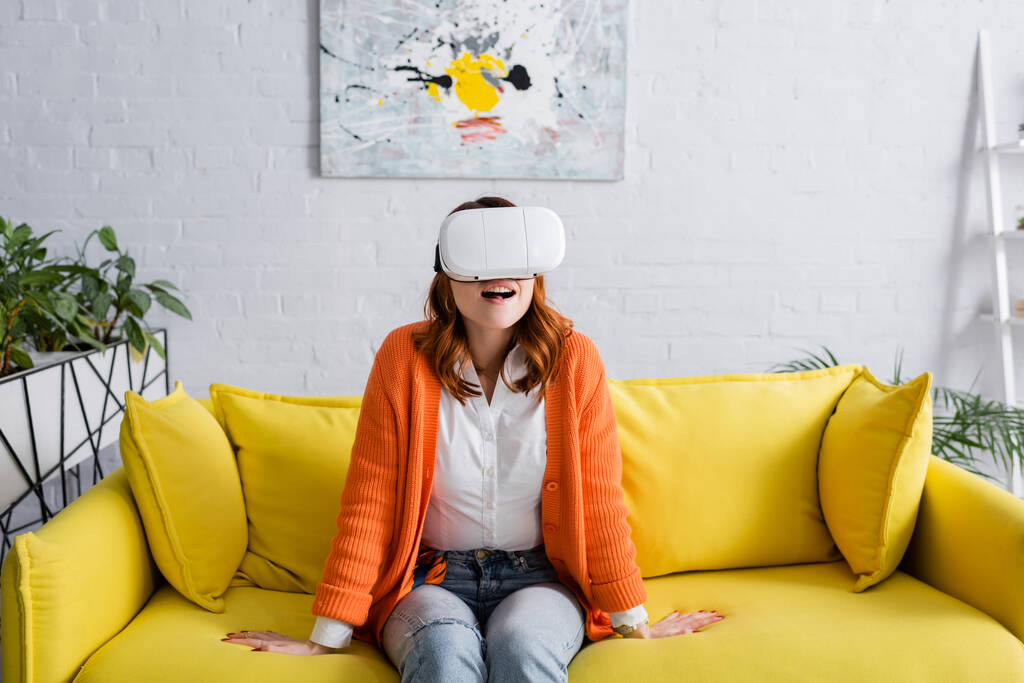 Oturma odasındaki sarı koltukta oturan VR kulaklıklı kadın hayrete düştü. - Fotoğraf, Görsel