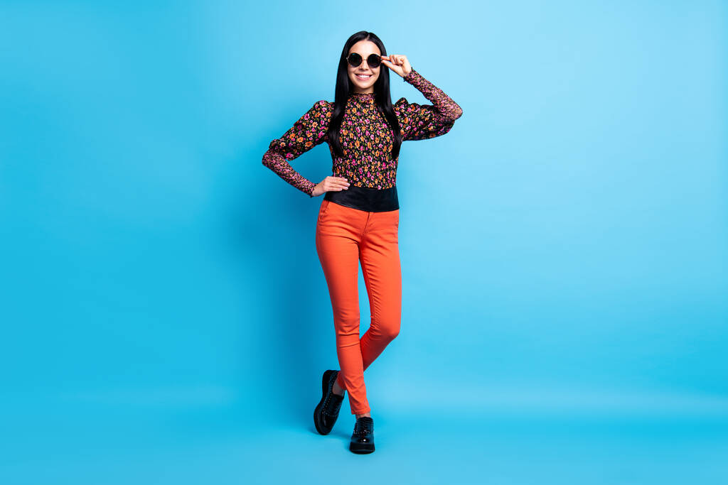 Foto van zelfverzekerde dame poseren look camera dragen retro specs bloem print shirt broek laarzen geïsoleerde blauwe kleur achtergrond - Foto, afbeelding