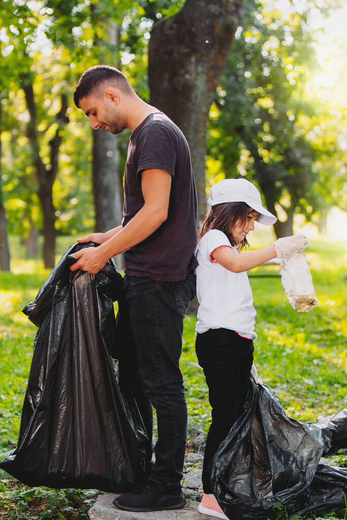 Täyspitkä kuva miehestä ja tytöstä seisomassa selkä selkää vasten pitelemässä roskapusseja ja keräämässä roskia. - Valokuva, kuva