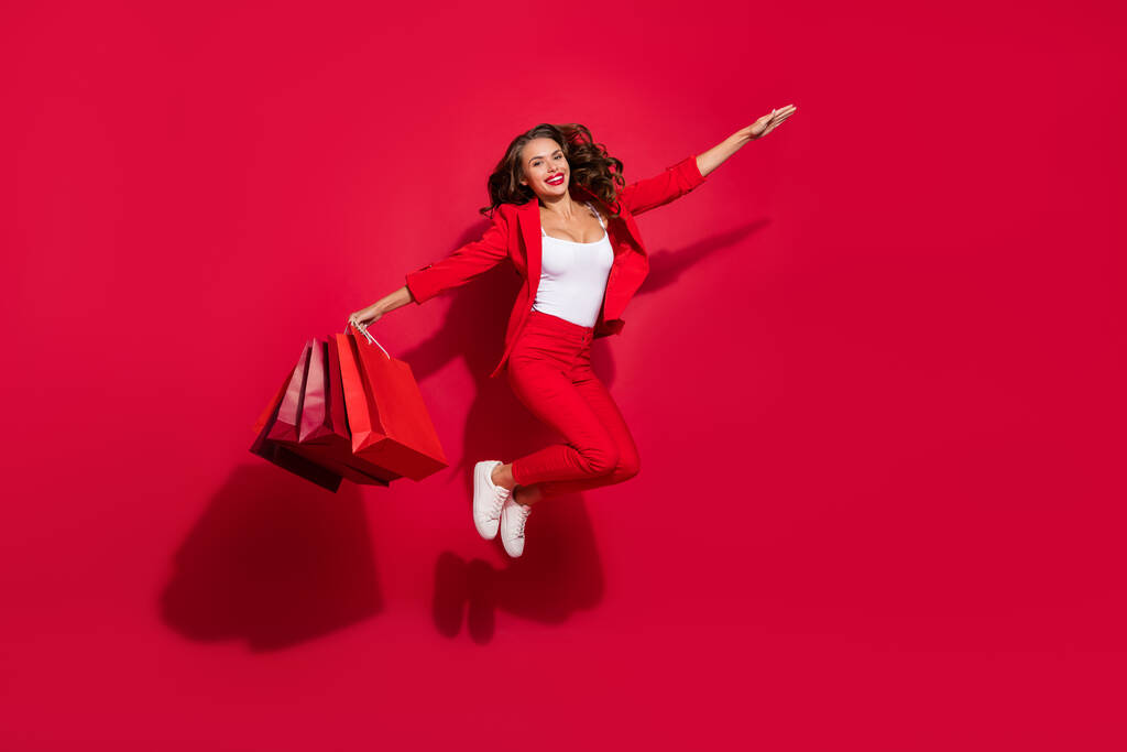Full body foto van onder de indruk brunette dame springen met tassen dragen pak schoenen geïsoleerd op rood kleur achtergrond. - Foto, afbeelding