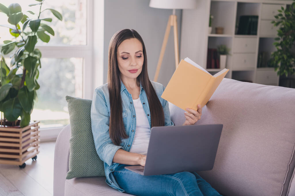 Portrait d'attrayant concentré femme aux cheveux longs assis sur divan en utilisant ordinateur portable livre de lecture préparant le projet à la maison à l'intérieur - Photo, image