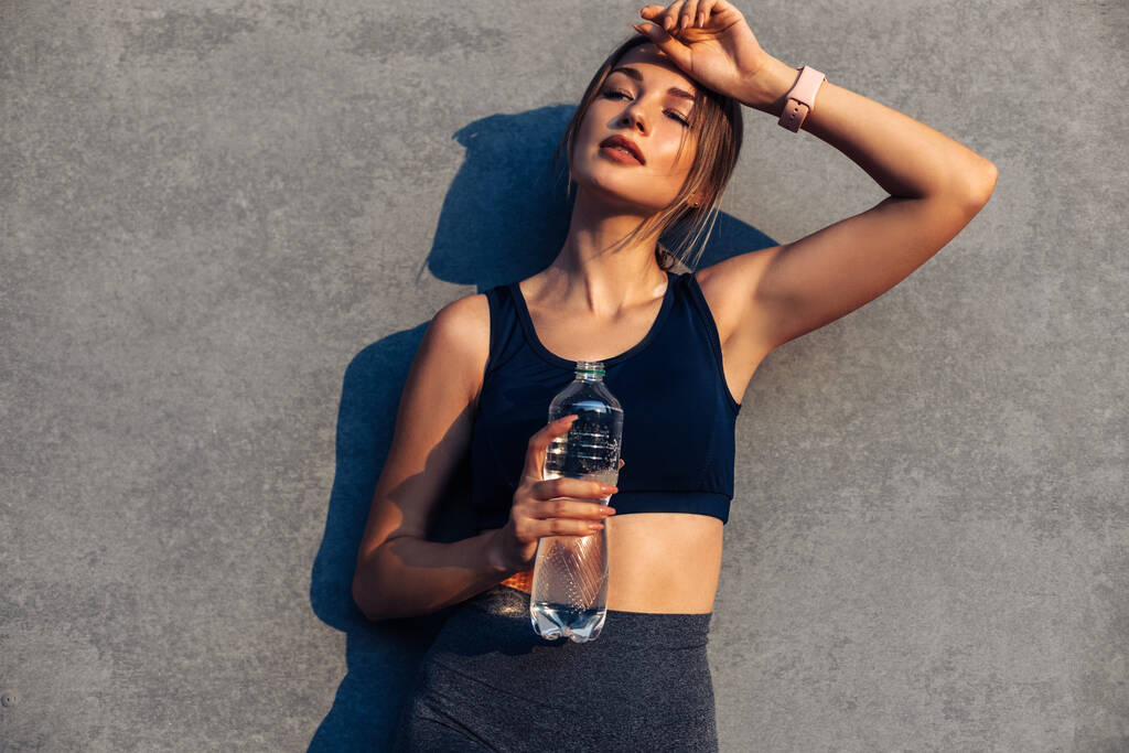 Втомлена спортивна жінка тримає пляшку води після ранкових тренувань, молода спортивна жінка стоїть на міській вулиці перед сірою стіною, після бігу, концепція здорового способу життя
 - Фото, зображення