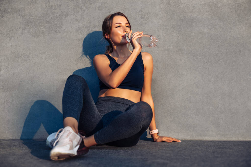 Moe atletische vrouw rusten na training buiten, Moe en uitgeput sportvrouw zitten op de vloer rusten en drinken van water, buiten in de buurt van de grijze stadsmuur - Foto, afbeelding