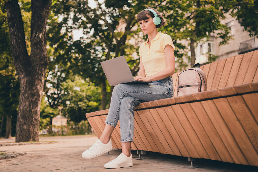 Full body foto di carino millennial bruna signora sedersi lavoro laptop indossare polo cuffie jeans scarpe da ginnastica fuori in città - Foto, immagini