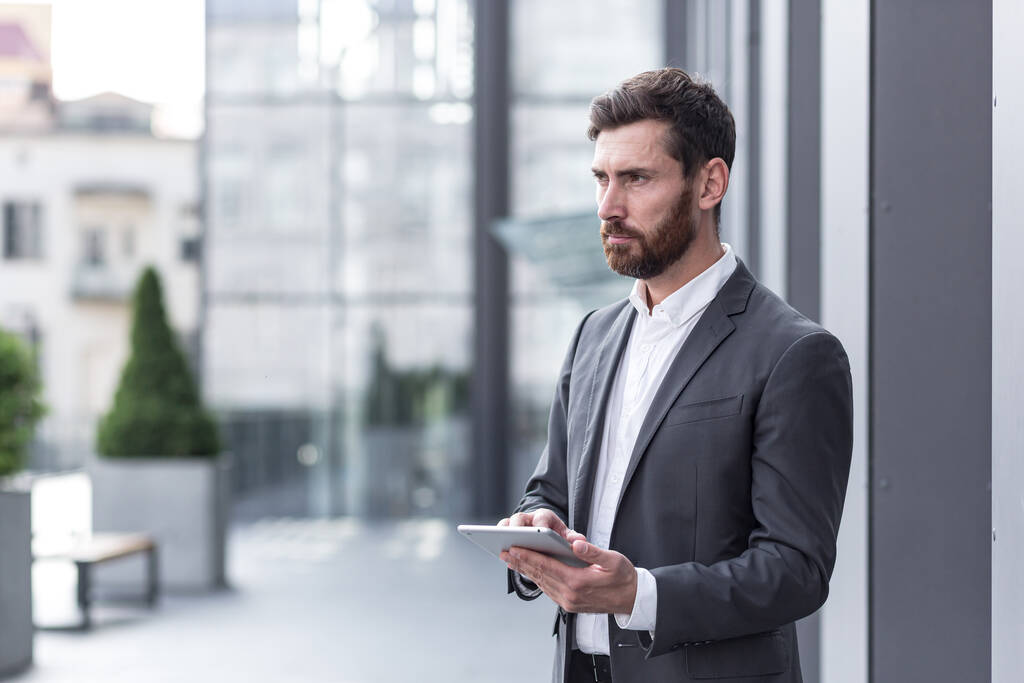 stílusos szakállas üzletember formális üzleti öltöny álló táblagéppel a kezében a háttér modern irodaház kívül. - Fotó, kép
