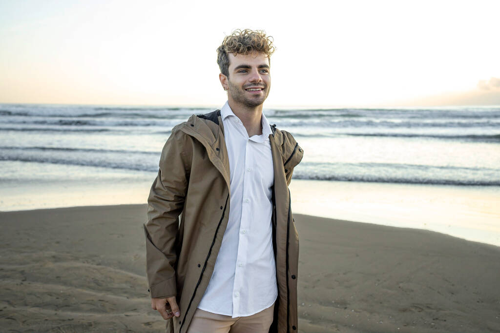 Un giovane attraente uomo caucasico dagli occhi azzurri con i capelli ricci in posa su una spiaggia - Foto, immagini