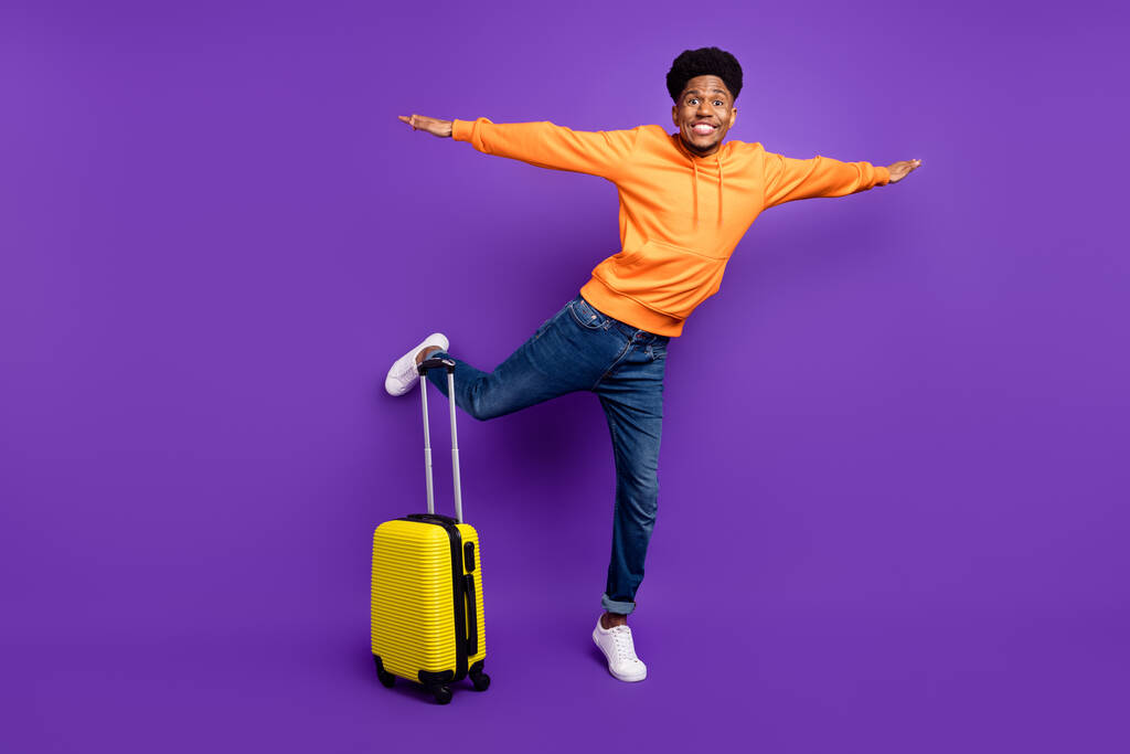 幸せなアフロアメリカのブルネットの男のフルサイズの写真はジーンズスーツケースの手の翼は、バイオレット色の背景に隔離されたフライ - 写真・画像