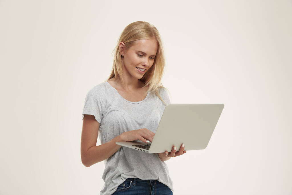 Jeune fille européenne souriante tenant et utilisant un ordinateur portable. Belle adolescente blonde aux yeux bleus porte un t-shirt blanc et un jean. Isolé sur fond blanc. Tournage studio - Photo, image
