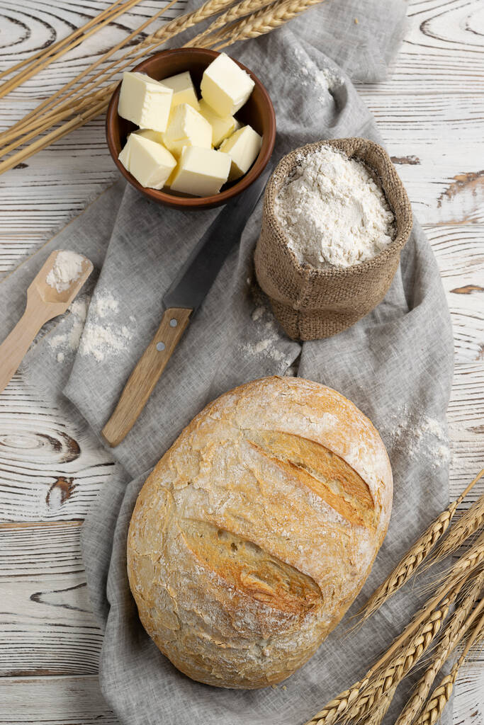 Una hogaza de pan blanco fragante con una corteza crujiente y harina en una bolsa de lona en una servilleta ligera. Orejas de trigo, un cuchillo y mantequilla en un tazón de barro sobre una mesa de madera clara. Vista superior - Foto, imagen