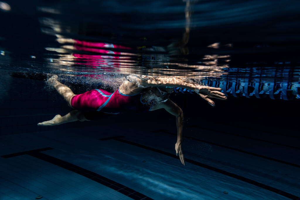 Eine Schwimmerin in Badekappe und Schwimmbrille trainiert im Schwimmbecken, drinnen. Unterwasser-Ansicht der Schwimmbewegungen Details. - Foto, Bild